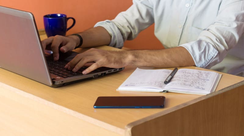 Foto de um jovem trabalhando em casa em um laptop sobre uma mesa junto com um laticínio e uma caneca de café