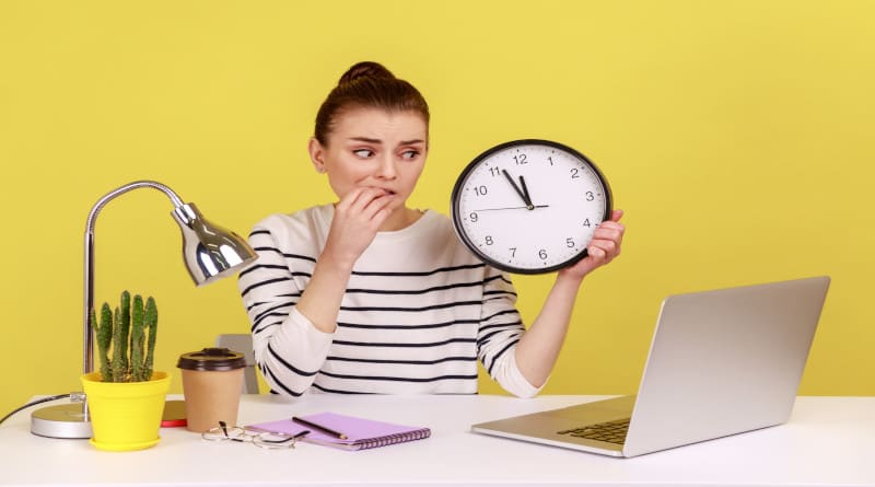 Foto de uma mulher infeliz, trabalhadora de escritório, roendo as unhas, segurando um relógio de parede, falando por videochamada no laptop
