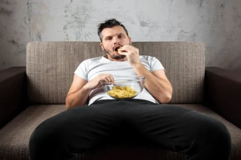Foto do cara de camisa deitado no sofá, comendo batatas fritas e assistindo um canal de esportes