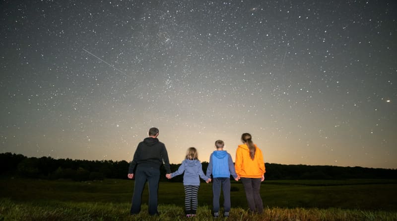 Foto de pais e filhos em pé no campo noturno observando o céu escuro com muitas estrelas brilhantes.