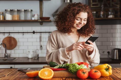 Foto de uma mulher caucasiana alegre segurando smartphone enquanto cozinha salada com legumes frescos no interior da cozinha