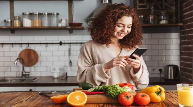Foto de uma mulher caucasiana alegre segurando smartphone enquanto cozinha salada com legumes frescos no interior da cozinha 