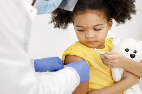 Médico masculino com bata médica branca cola um curativo após a vacinação. Foto recortada do rosto de uma garota. Menina vestindo camiseta amarela e segurando um brinquedo de urso nas mãos.
